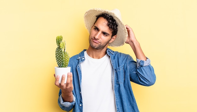 Un bell'uomo ispanico che si sente perplesso e confuso, graffia la testa dell'agricoltore e il concetto di cactus