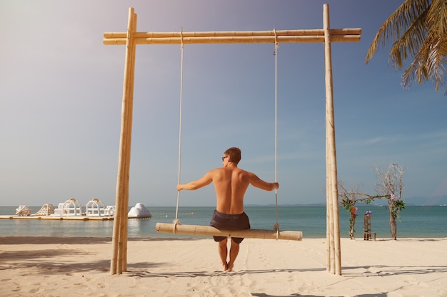 Un bell'uomo oscilla sulla spiaggia di sabbia e si gode il paesaggio sotto le palme verdi contro la vista sul retro del cielo blu