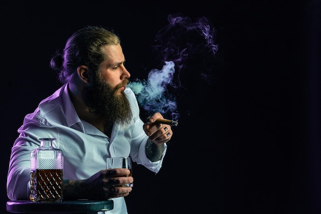 Un bell'uomo seduto con la barba che fuma un sigaro tiene un bicchiere di whisky vestito con una camicia bianca in studio