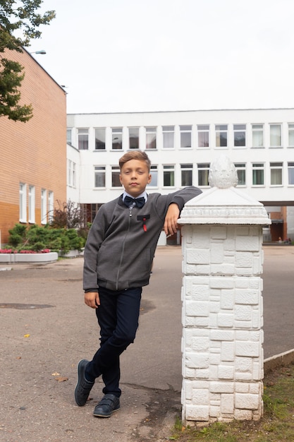 Un bellissimo bambino di otto anni in uniforme scolastica, pantaloni neri e camicia bianca con farfallino posa nel cortile della scuola.