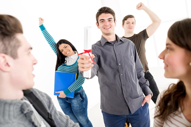 Un bellissimo giovane studente sorridente con diploma. Dietro di lui c'è un gruppo felice di suoi amici con le braccia alzate a pugno in aria.