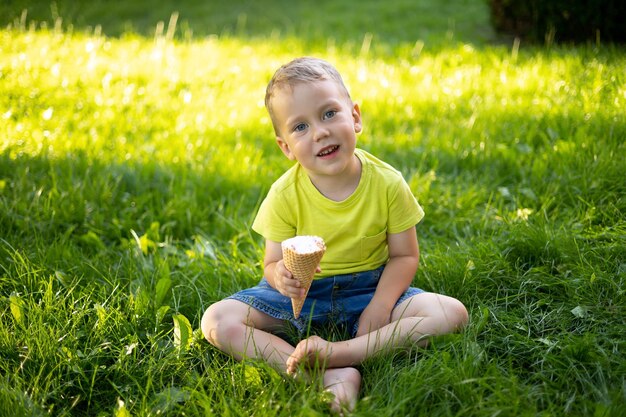 Un bellissimo ragazzino con gli occhi azzurri e i capelli biondi mangia il gelato nel parco si siede sull'erba verde sorride gioisce