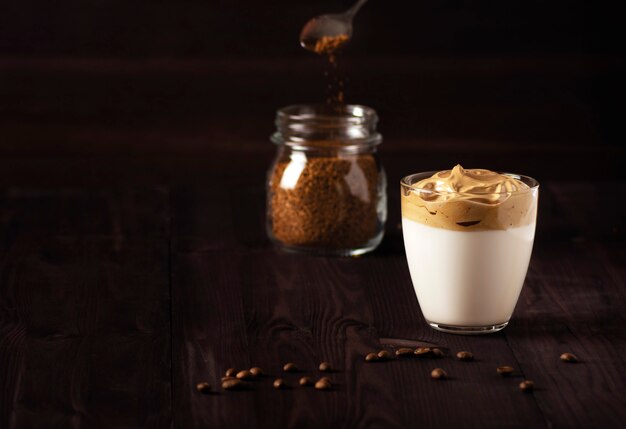 Un bicchiere di caffè Dalgona e una lattina di caffè istantaneo stanno su un tavolo di legno scuro.