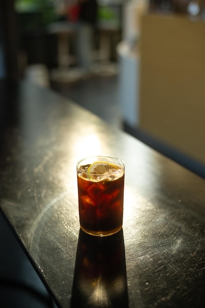 Un bicchiere di caffè nero si trova sul bancone di un bar con una luce gialla che brilla sul vetro.