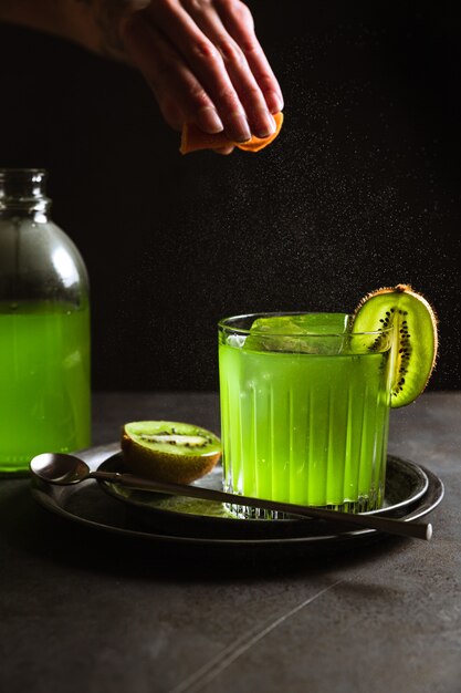Un bicchiere rock con una bevanda verde rinfrescante e un cubetto di ghiaccio all'interno, guarnito con kiwi, una bottiglia di bevanda verde, un cucchiaio da bar. Colpo di espressione di olio di agrumi.