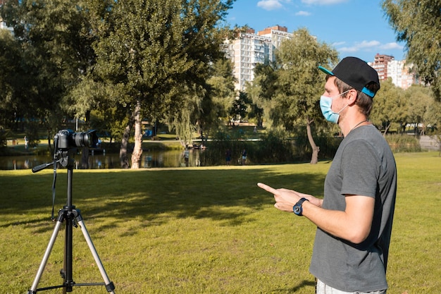 Un blogger in una maschera medica protettiva registra un video blog su una fotocamera digitale nel parco.