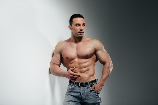 Un bodybuilder giovane atleta posa in topless in jeans vicino al muro. Sport.