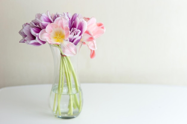 Un bouquet aperto di tulipani rosa e viola si trova in un vaso di vetro trasparente su un tavolo bianco