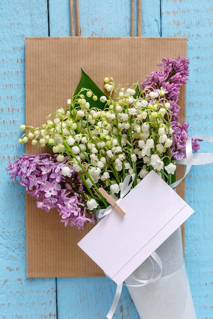 Un bouquet di mughetti con lillà in un sacchetto di carta su un blu di legno con un biglietto da visita allegato