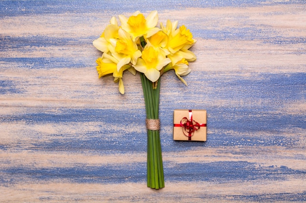 Un bouquet di narcisi, un regalo con un nastro rosso su uno sfondo di legno. Fiori gialli di primavera. Vernice peeling blu. Design piatto laici, vista dall'alto.