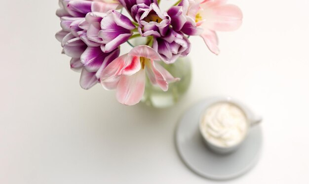 Un bouquet di tulipani rosa e viola aperti e una tazza bianca con caffè con latte su un tavolo bianco