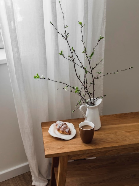 Un caffè accogliente e estetico una tazza di caffè un croissant una brocca con rami di primavera su una panchina di legno accanto alla finestra