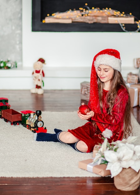 Un cappello allegro sveglio della Santa della ragazza vicino all'albero di Natale. Tiene la palla di Natale rossa.