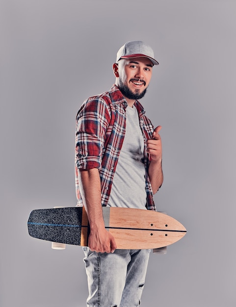 Un carismatico giovane hipster con la barba in camicia a quadri e berretto punta il dito contro qualcuno con un longboard su uno sfondo grigio in studio.