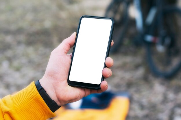 Un ciclista uomo tiene un modello di uno smartphone con uno schermo bianco tra le mani sullo sfondo di una bicicletta in natura.