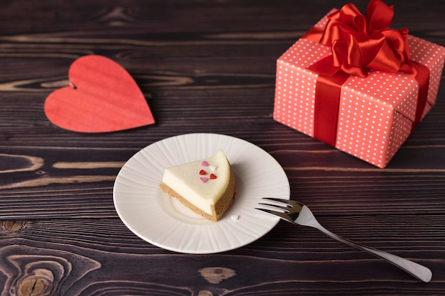 Un classico pezzo di cheesecake su un piatto bianco con un cuore rosso e un regalo su un tavolo di legno Fuoco selettivo