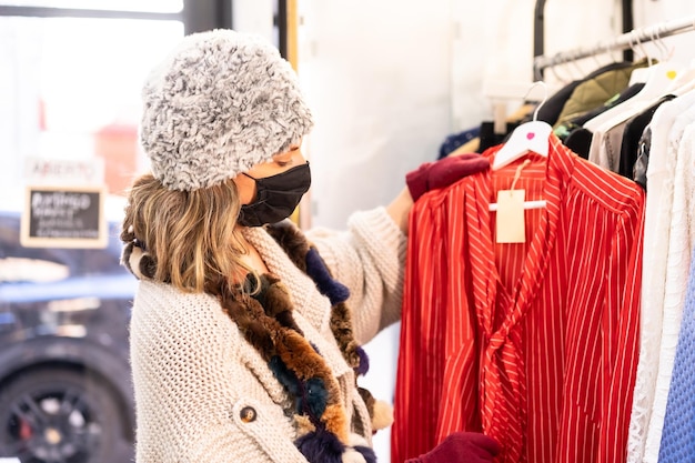 Un cliente con una maschera protettiva che guarda i vestiti della nuova collezione su un appendiabiti, un negozio di abbigliamento di seconda mano e sostenibile, coronavirus, covid-19