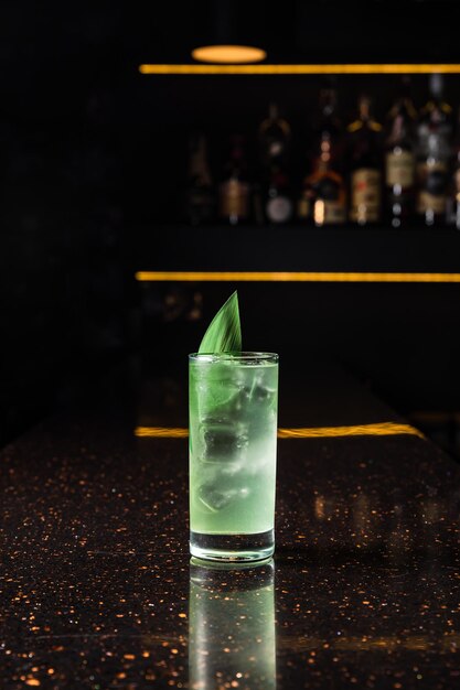 Un cocktail alcolico verde chiaro in un bicchiere highball con ghiaccio, guarnito con una foglia di bambù, al bancone del bar, sfondo scuro