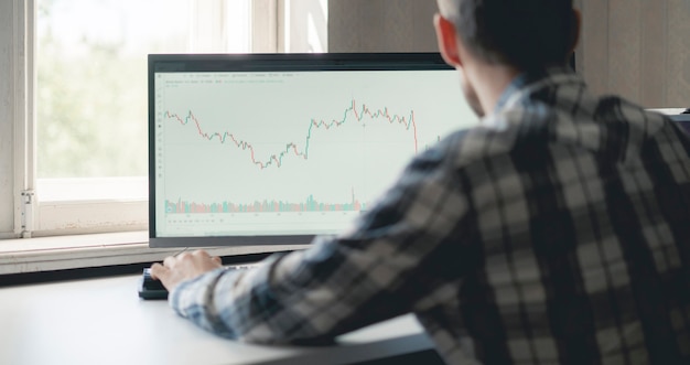 Un commerciante maschio che guarda il grafico azionario sul grande schermo del computer a casa, analisi del grafico