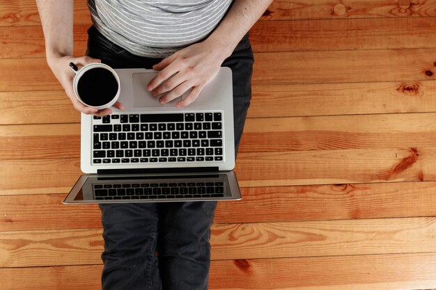 Un computer portatile e una tazza di caffè nelle mani di un uomo seduto su un pavimento di legno.
