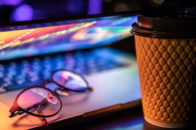 Un computer portatile mezzo chiuso al buio con una tazza di caffè e bicchieri colorati