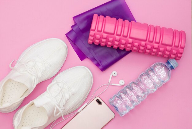 Un concetto di fitness con una bottiglia d'acqua, scarpe da ginnastica, un telefono con le cuffie, un elastico viola per il fitness, un rullo rosa su uno sfondo rosa. Copia spazio.