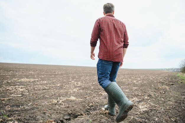 Un contadino con gli stivali lavora con il suo tablet in un campo seminato in primavera Un agronomo cammina per terra valutando un campo arato in autunno Agricoltura Tecnologie agricole intelligenti