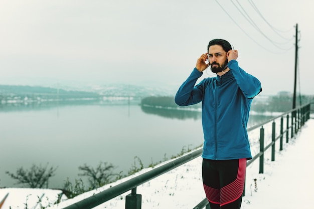 Un corridore maschio con le cuffie sulle orecchie che si prende una pausa nel luogo pubblico durante l'allenamento invernale fuori accanto al fiume. Copia spazio.