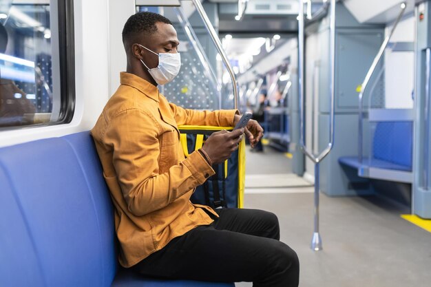 Un corriere afroamericano con una maschera protettiva tiene in mano un telefono cellulare mentre viaggia in un vagone della metropolitana con uno zaino giallo