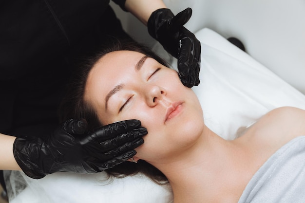 Un cosmetologo fa un massaggio facciale cosmetico per il viso di una giovane donna rilassata in una spa di bellezza Massaggio per la cura del viso concetto di cosmetologia per la cura della pelle