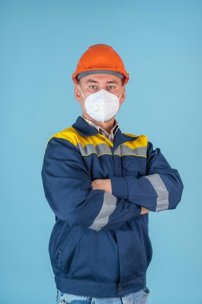 Un costruttore professionista con un elmetto arancione e una maschera protettiva in piedi con le braccia incrociate su uno sfondo blu.