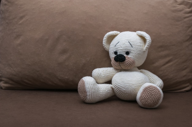 Un cucciolo di orso bianco lavorato a maglia su un morbido divano marrone. Bellissimo giocattolo lavorato a maglia.