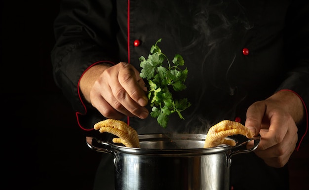 Un cuoco in uniforme nero aggiunge il prezzemolo a una padella di zampe di pollo bollenti Prepara una deliziosa zuppa o un contorno grasso in cucina Concetto di cottura con pollo o gallo su sfondo nero