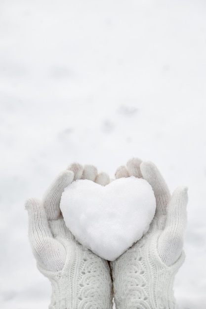 Un cuore di neve nelle mani che indossa guanti a maglia sullo sfondo di un manto nevoso