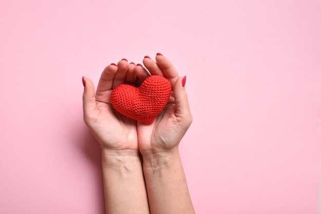 Un cuore rosso lavorato a maglia nelle mani di una ragazza con una bella manicure su sfondo rosa Amore Donazione di organi Gravidanza San Valentino Spazio copia Vista dall'alto piatta