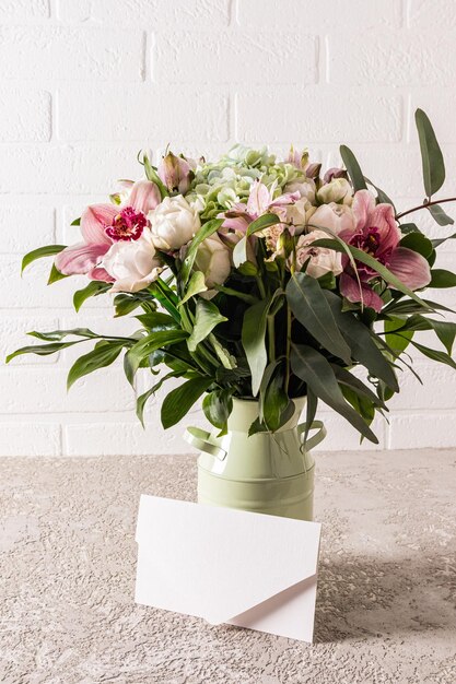 Un delicato bouquet di fiori freschi di astromeria in un vaso di metallo decorativo e una busta vuota bianca su un tavolo grigio di fronte a un muro di mattoni bianchi