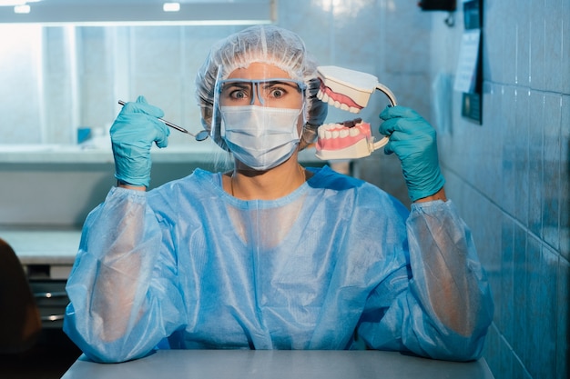 Un dentista che indossa guanti blu e una maschera tiene un modello dentale della mascella superiore e inferiore e uno specchio dentale.