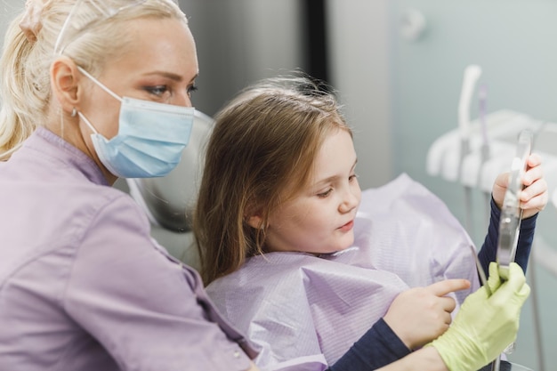 Un dentista femminile e una bambina carina tengono uno specchio e guardano i suoi denti dopo l'esame dentale presso l'ufficio del dentista.