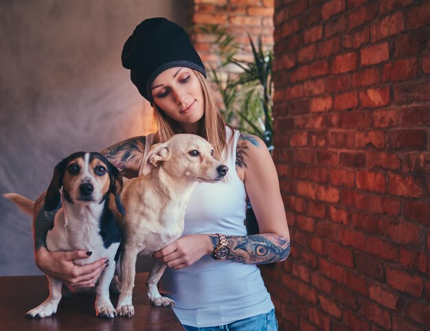 Un'elegante donna bionda tatuata in maglietta e jeans abbraccia due simpatici cani.