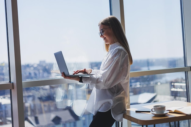 Un'elegante donna d'affari con gli occhiali per la correzione della vista tiene in mano un laptop moderno in piedi vicino a una finestra a parete intera in un ufficio moderno