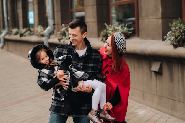 Un'elegante famiglia di tre persone sta passeggiando per la città in autunno e papà tiene in braccio sua figlia