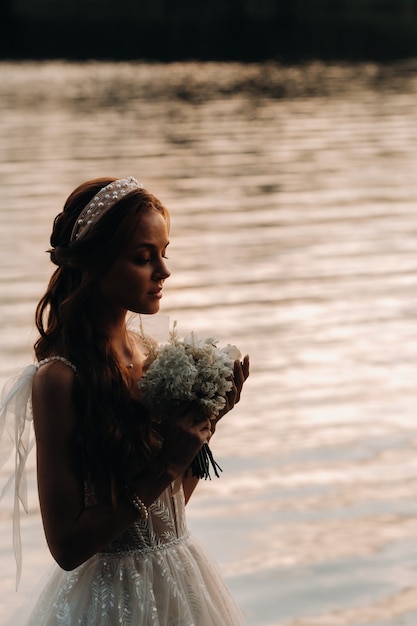 Un'elegante sposa in abito bianco e guanti si trova in riva al fiume nel parco con un bouquet, godendosi la natura al tramonto. Un modello in abito da sposa e guanti in un parco naturale. Bielorussia