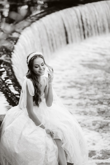 Un'elegante sposa in abito bianco, guanti e piedi nudi è seduta vicino a una cascata nel parco godendosi la natura. Un modello in abito da sposa e guanti in un parco naturale. Bielorussia