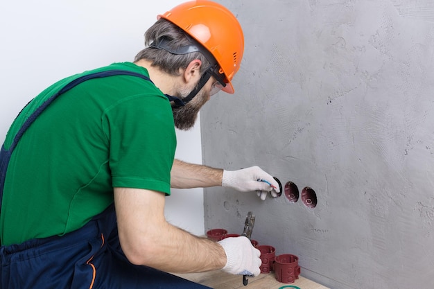 Un elettricista installa le prese nell'appartamento. Un ragazzo con un casco e una tuta arancione fa l'impianto elettrico in casa.