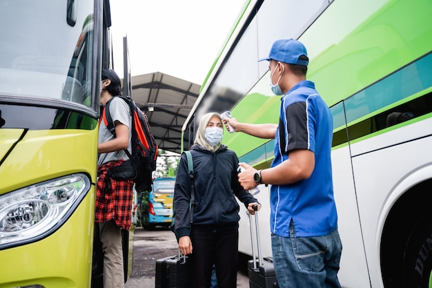 Un equipaggio di autobus in uniforme e cappello con una pistola termica ispeziona il passeggero di una donna con velo e maschera prima di salire sull'autobus