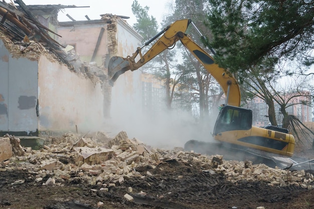 Un'escavatore e altre attrezzature pesanti di costruzione lavorano sul cantiere Un'excavatore che usa un secchio demolisce le pareti di una vecchia casa fatiscente