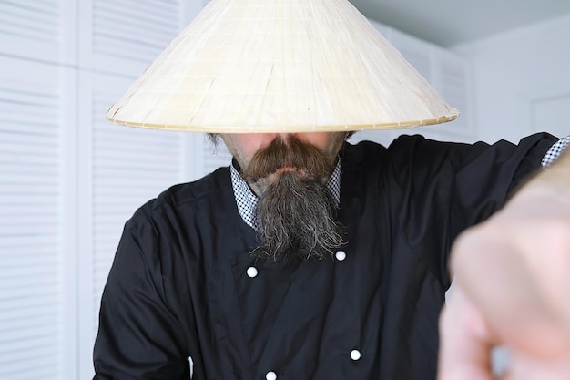 Un'esilarante parodia di un uomo asiatico con un cappello vietnamita con la barba. Ritratto. Chef asiatico del caffè.