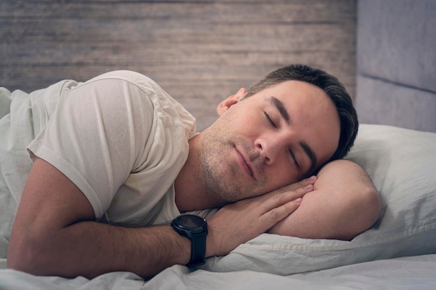 Un'espressione soddisfatta sul suo viso giovane sta riposando a letto con uno smartwatch sulle mani dormendo un bell'uomo