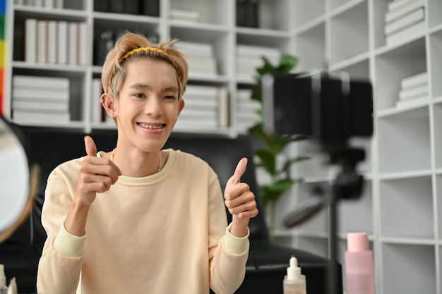 Un felice blogger gay asiatico sta mostrando i suoi pollici alla telecamera e sta registrando un video