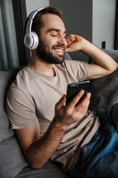 un felice giovane uomo positivo al chiuso a casa sul divano ascoltando musica con le cuffie utilizzando il telefono cellulare.